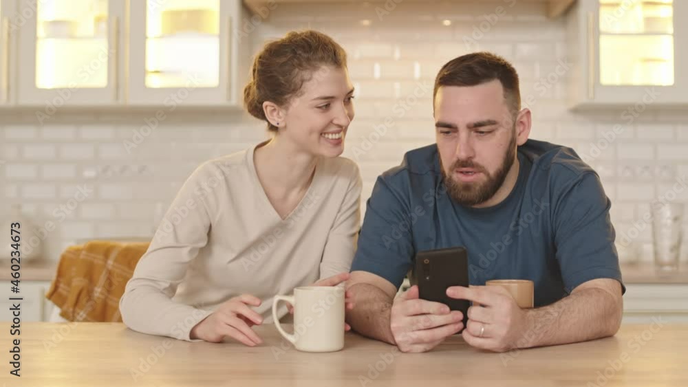 Waist-up of bearded Caucasian man sitting at table in kitchen, using smartphone, his beautiful wife holding coffee mug, giving him present, smiling Waist-up of bearded Caucasian man sitting at table in kitchen, using smartphone, his beautiful wife holding coffee mug, giving him present, smiling