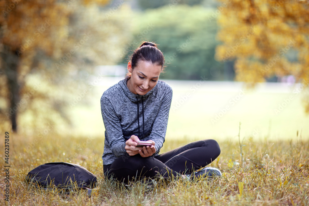 Young woman with happy face dressed in outdoor sportswear sitting on the grass in park and checks messages on smartphone Young woman with happy face dressed in outdoor sportswear sitting on the grass in park and checks messages on smartphone