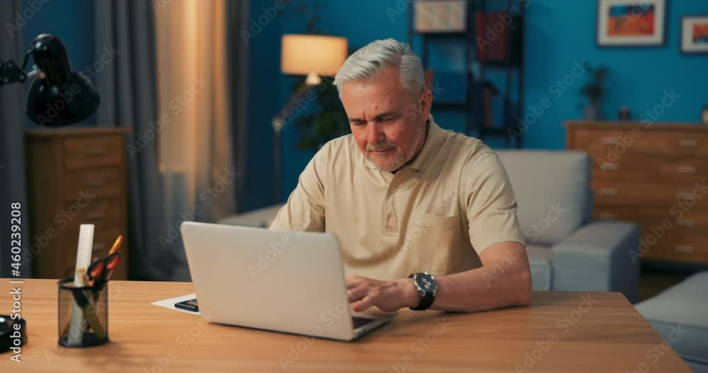 Elderly man sitting at desk with laptop in the evening. Grandfather talks to grandchildren online. A smiling and happy senior talks through the camera of his laptop. An elderly man chats online. Elderly man sitting at desk with laptop in the evening. Grandfather talks to grandchildren online. A smiling and happy senior talks through the camera of his laptop. An elderly man chats online.