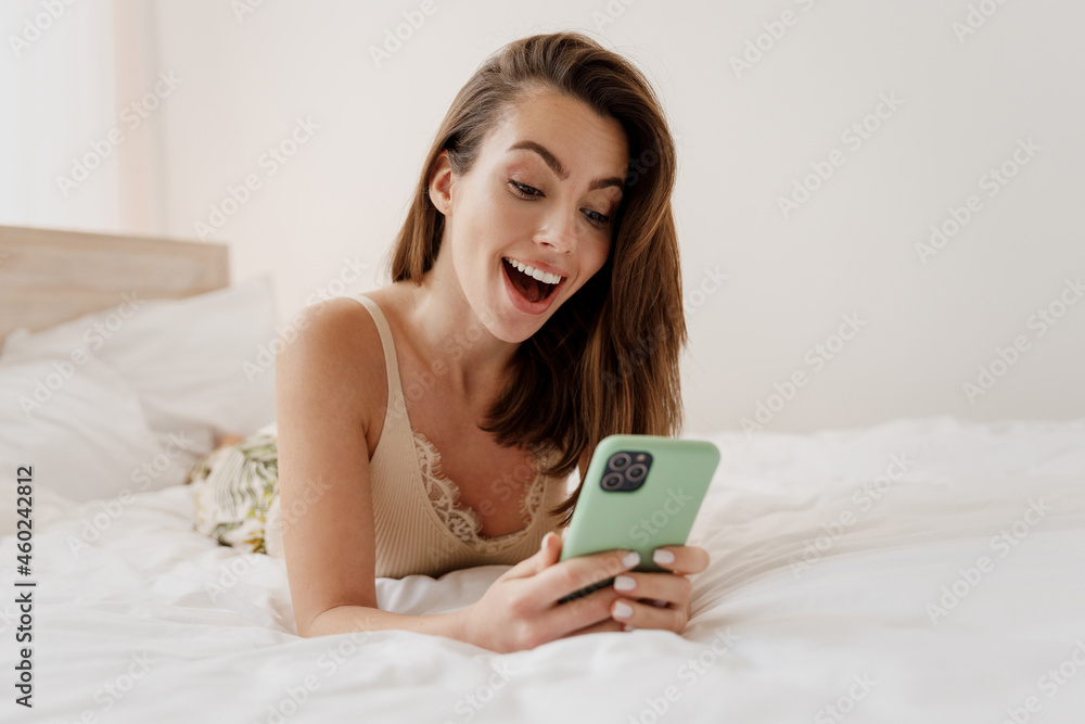 Shocked surprised fun young woman in casual casual clothes lying on her stomach hold in hand use mobile cell phone keep mouth wide open rest relax spend time in bedroom lounge home in own room house.