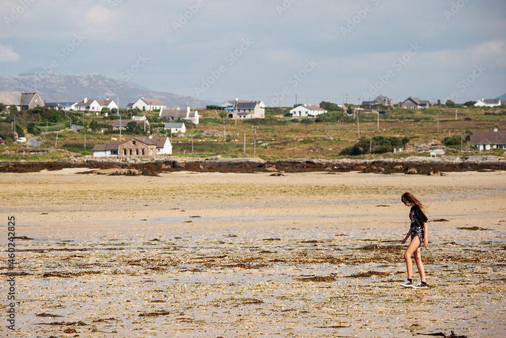 Teenager girl walking on a sandy beach at low tide. Houses and mountains in the background. Warm sunny day. Outdoor activity. Copy space