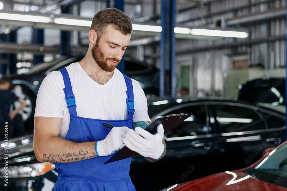 Young serious car mechanic man in denim blue overalls white t-shirt gloves estimate hold clipboard with papers document work in light modern vehicle repair shop workshop indoors. Tattoo translate fun. Young serious car mechanic man in denim blue overalls white t-shirt gloves estimate hold clipboard with papers document work in light modern vehicle repair shop workshop indoors. Tattoo translate fun.