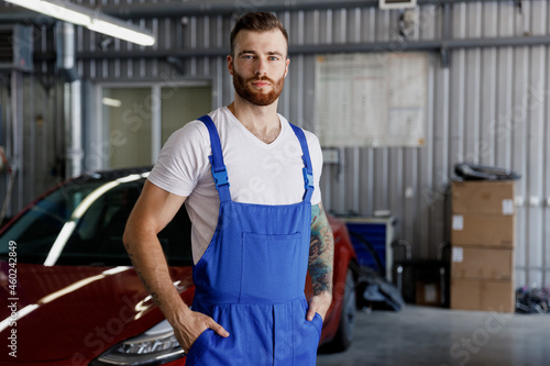 Strong serious young male professional technician car mechanic man in denim blue overalls white t-shirt stand near red automobile look camera work in light modern vehicle repair shop workshop indoors.