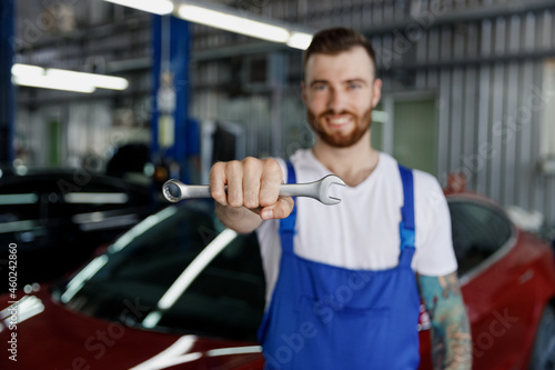 Smiling fun happy young male professional technician car mechanic man in denim blue overalls white t-shirt hold give wrench key tool work in modern vehicle repair shop workshop inside Focus on spanner