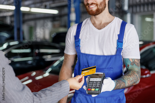 Cropped young happy professional car mechanic man in blue overalls gloves hold payment terminal fow paying with credit card by female owner driver woman work in vehicle repair shop workshop indoors photo