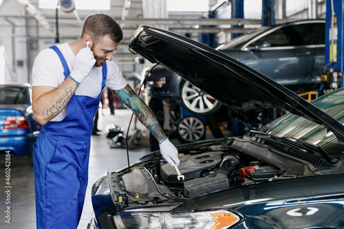 Young technician car mechanic man in blue overalls white t-shirt gloves listen music in earphones fix problem with raised hood bonnet hold spanner work in modern vehicle repair shop workshop indoor