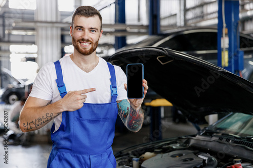 Young technician car mechanic man in overalls white t-shirt use hold point finger on mobile cell phone blank screen area fix problem with raised hood bonnet work in vehicle repair shop workshop inside