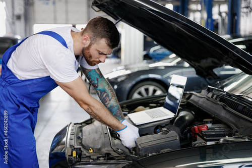 Young repairman professional technician car mechanic man in blue overalls t-shirt using laptop pc computer make diagnostics fix problem with raised hood work in vehicle repair shop workshop indoor