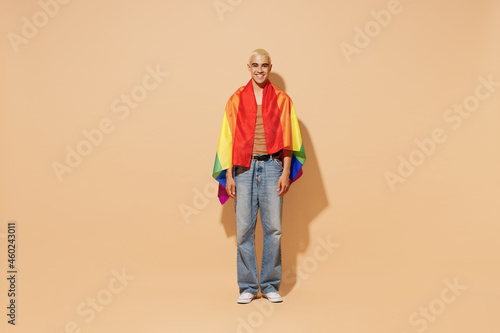 Full body young activist smiling happy fun cool blond latin gay man with make up in beige tank shirt wrapped rainbow flag isolated on plain light ocher background studio People lgbt lifestyle concept