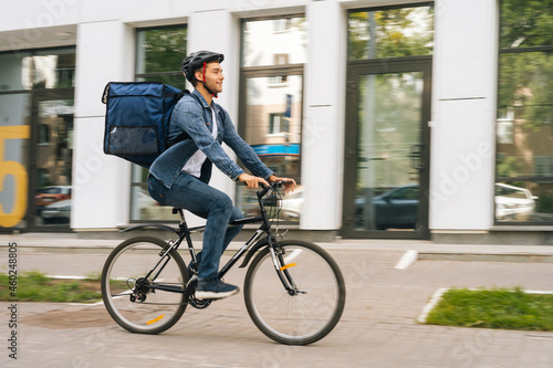 Side view of handsome young delivery man with thermo backpack riding bicycle in city street on blurred background of office building. Courier male in protective helmet delivery food to client