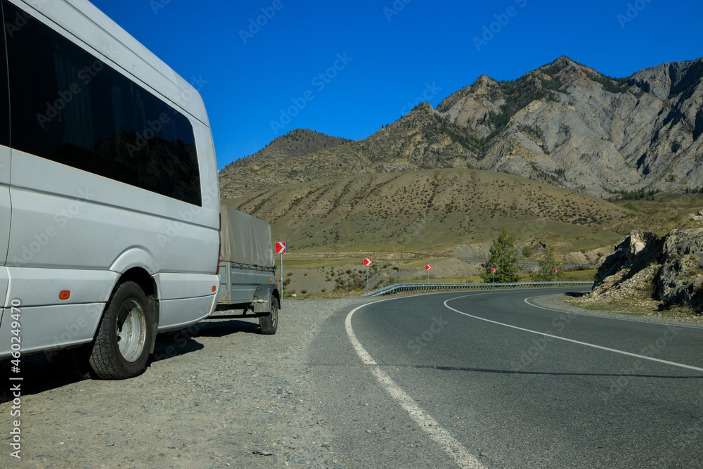a white minibus with a trailer is standing on the side of the highway among the mountains. The road turns around a bend. Against the background of steep high mountains and clear blue sky