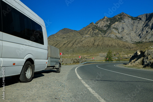 a white minibus with a trailer is standing on the side of the highway among the mountains. The road turns around a bend. Against the background of steep high mountains and clear blue sky