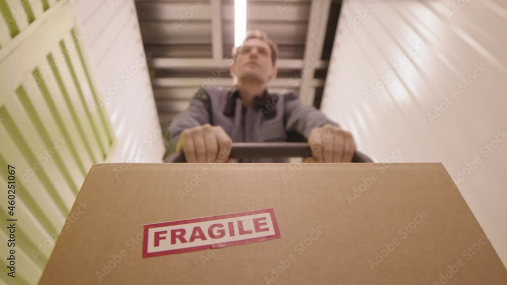 Low angle tracking of blurred male worker pushing trolley with cardboard box with sticker saying fragile on it in hallway of warehouse
