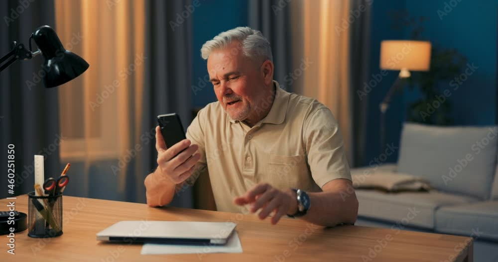 Older man laughs into phone webcam. Elderly executive is video conferencing on smartphone. Senior men is chatting online. The grey-haired guy is gesturing with hands while talking on the telephone Older man laughs into phone webcam. Elderly executive is video conferencing on smartphone. Senior men is chatting online. The grey-haired guy is gesturing with hands while talking on the telephone