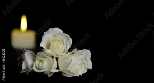 Beautiful White roses with a burning candle on the dark background. Funeral flower and candle on table against black background with copy space. Funeral symbol. Mood and Condolence card concept.