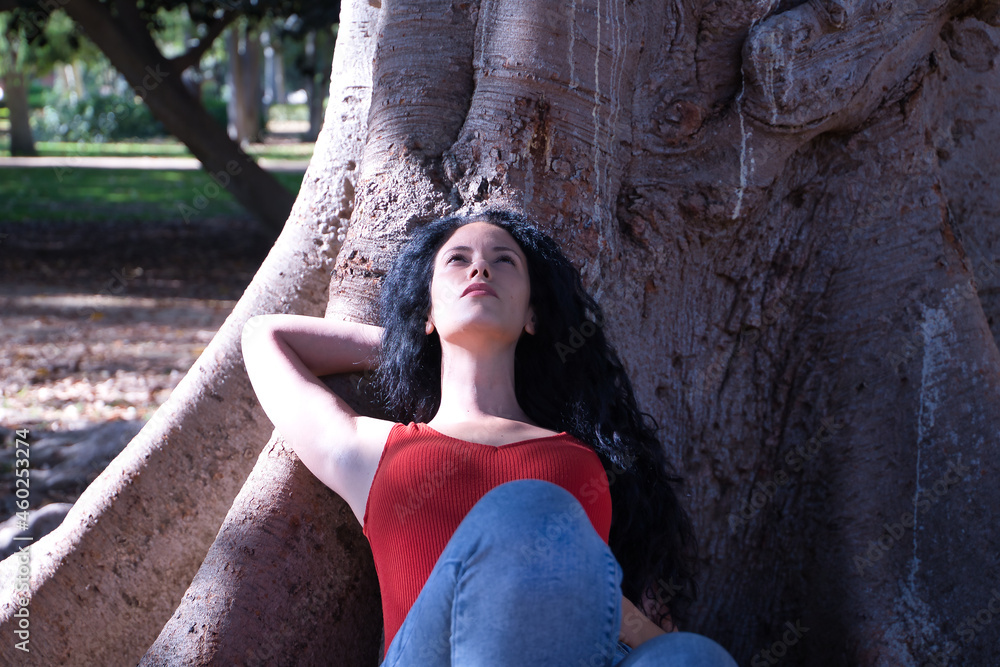 Middle-aged adult Hispanic woman with black curly hair, lying at the root of a tree, looking thoughtfully up at the sky. Concept thoughts, escape, nature.