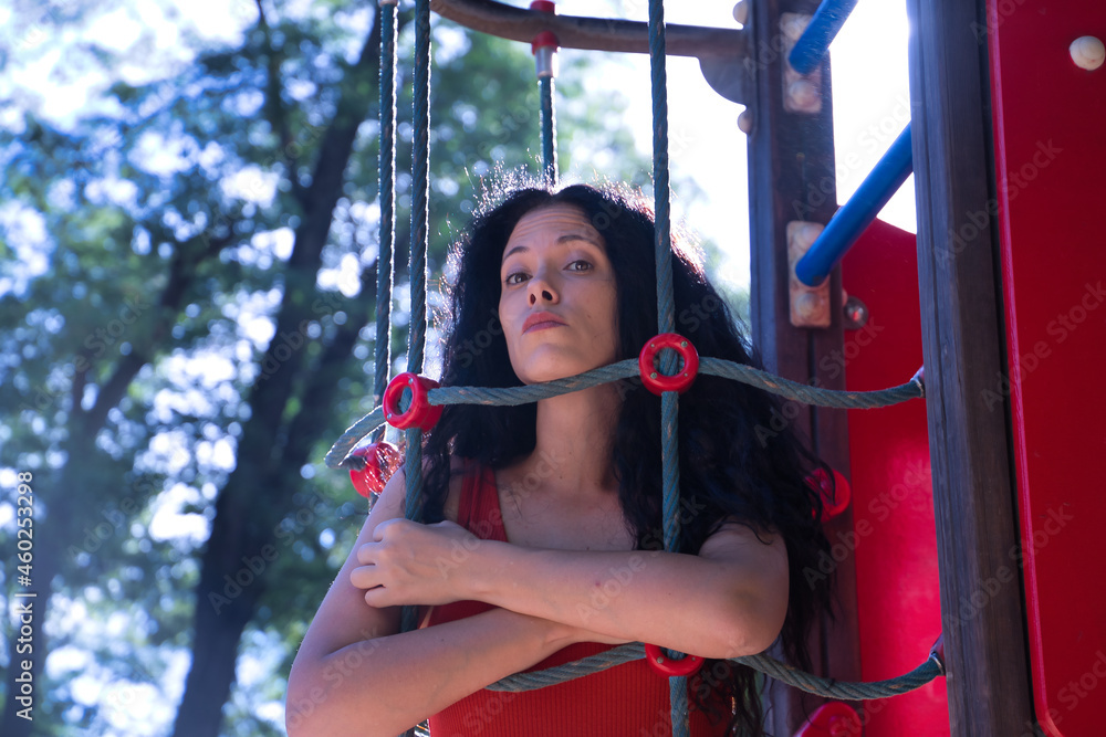 Obraz premium Middle-aged adult Hispanic woman with black curly hair, hugging the ropes of a climbing apparatus, shyly. Concept games, shyness, looks, sensations.