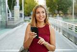 Smiling young woman is walking in the street with phone. She is holding gadget in hand and looking away.