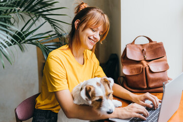 Diligent smiling female freelancer typing on the computer with her cute jack russell terrier dog at workplace at home office or cafe. People and dog friendship concept.