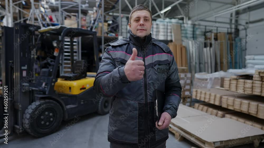 Positive confident man in uniform showing thumb up smiling looking at camera. Caucasian warehouse worker posing in storage site in slow motion. Occupation and lifestyle concept Positive confident man in uniform showing thumb up smiling looking at camera. Caucasian warehouse worker posing in storage site in slow motion. Occupation and lifestyle concept