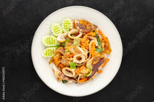 Pad See Ew Pla Muek, Thai food, stir fried soy sauce noodle with squid, egg, lettuce, carrot served with cucumber on dark tone texture background, top view