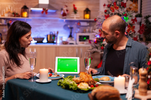 Cheerful family enjoying xmas dinner in x-mas decorated kitchen celebrating christmas holiday. Green screen mock up chroma key tablet computer with isolated display standing on table