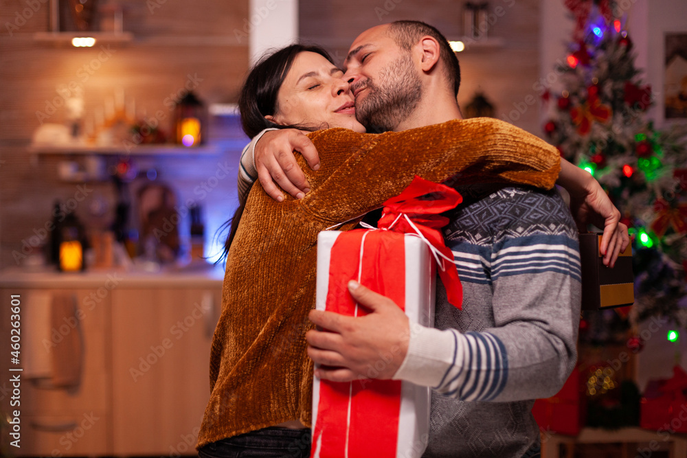 Happy family enjoying spending time together during christmas holiday holding xmas decorated present gift with ribbon on it. Excited married couple hugging celebrating winter festive season