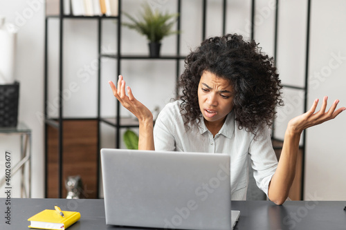 Confused African-American businesswoman in casual shirt sitting at the desk in front of laptop and spreading hands, mixed-race female office employee has issue with project, computer error photo