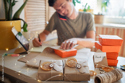 Young millennial worker packing Christmas gift boxes for clients, family small business. New Year celebration, distant work and home office, indoor lifestyle photo
