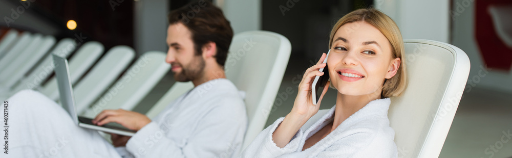 Happy woman in bathrobe talking on smartphone near blurred boyfriend with laptop in spa center, banner Happy woman in bathrobe talking on smartphone near blurred boyfriend with laptop in spa center, banner