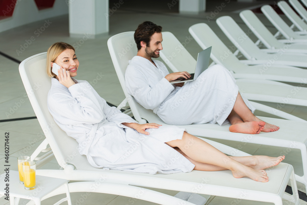 Fototapeta premium Cheerful woman talking on smartphone near orange juice and boyfriend with laptop in spa center