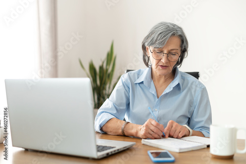 Serious mature senior female office worker business lady listening to an online webinar on laptop, taking notes in notebook, working on freelance project at home, remote work, learning at home concept