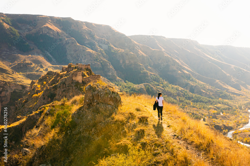 Female woman walks up the hill towards Tmogvi fortress surrounded by golden autumn nature and dramatic landscape in caucasus.