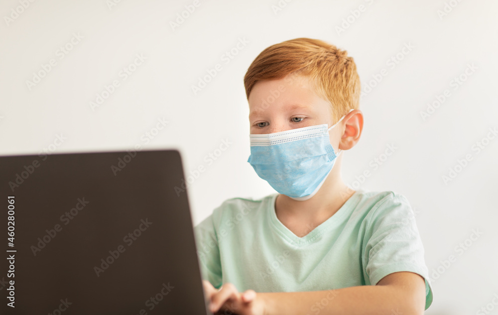 Redhead boy in face mask using laptop, home-schooling concept Redhead boy in face mask using laptop, home-schooling concept