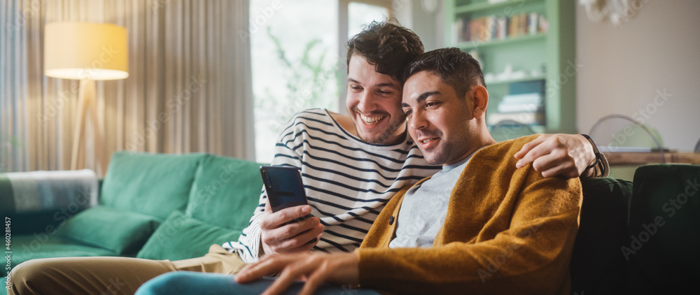 Obraz premium Portrait of Gentle Gay Couple Using Smartphone, while Sitting on a Couch in Cozy Stylish Apartment. Adult Boyfriends Online Shopping on Internet, Watching Funny Videos on Streaming Service.