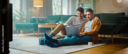 Handsome Gay Couple Using Laptop Computer, while Sitting on a Living Room Floor in Cozy Stylish Apartment. Adult Boyfriends Online Shopping on Internet, Watching Funny Videos on Streaming Service.