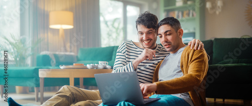 Handsome Gay Couple Using Laptop Computer, while Sitting on a Living Room Floor in Cozy Stylish Apartment. Adult Boyfriends Online Shopping on Internet, Watching Funny Videos on Streaming Service.