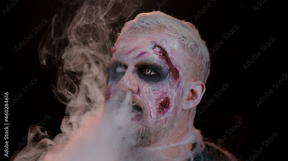 Creepy man with bloody scars face, Halloween stylish zombie make-up. Scary wounded undead guy blows smoke from nose, smiles terribly. Voodoo rituals. Thematic party. Sinister beast, monster, vampire