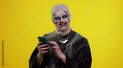 Frightening man with Halloween zombie bloody wounded makeup using mobile phone typing new post on web sms message, browsing, addiction of social networks. Sinister undead guy on yellow wall background © Andrii Iemelianenko