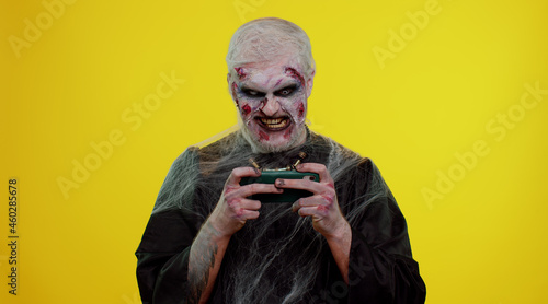 Sinister man with horrible scary Halloween zombie makeup enthusiastically playing racing drive video games on mobile phone. Dead guy with wounded bloody scars face isolated against yellow background © Andrii Iemelianenko