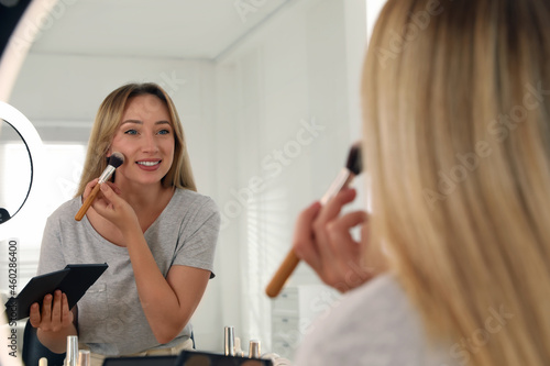 Beautiful young woman applying highlighter with brush near mirror indoors