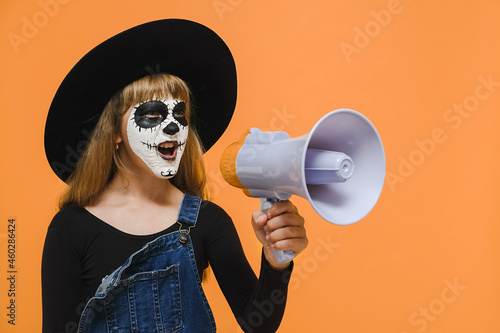 Portrait of spooky girl kid with Halloween makeup mask screaming in megaphone looking aside, wears big black hat, posing isolated over orange color background wall in studio. Party holiday concept
