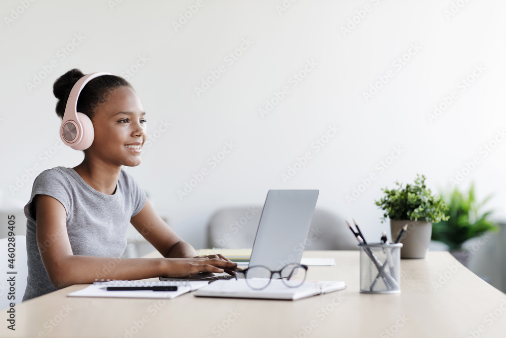 Happy teen afro american girl in headphones watch video lesson remotely at laptop in living room interior Happy teen afro american girl in headphones watch video lesson remotely at laptop in living room interior