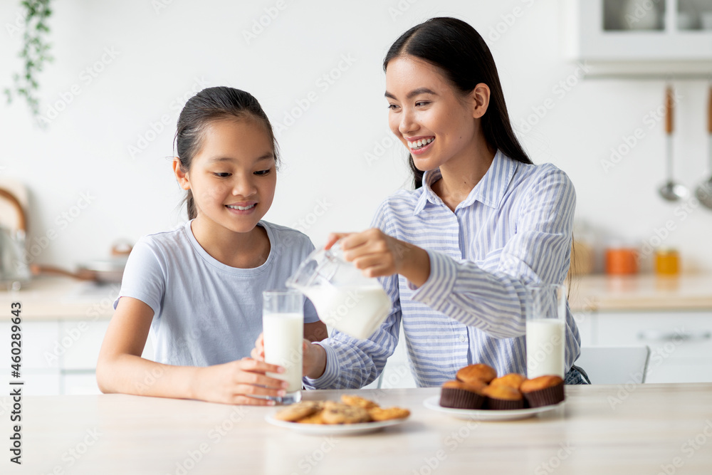 Happy asian mom and daughter eating snacks and drinking milk in kitchen interior, having a bite together Happy asian mom and daughter eating snacks and drinking milk in kitchen interior, having a bite together