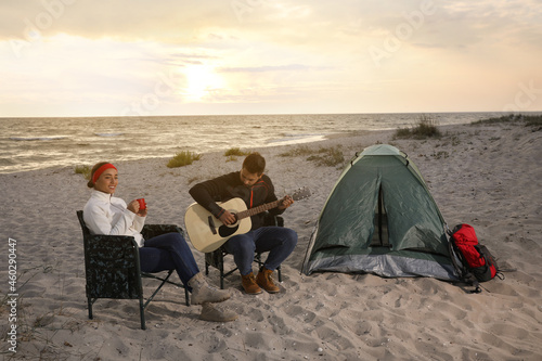 Young man playing guitar to his beloved girlfriend near camping tent on beach