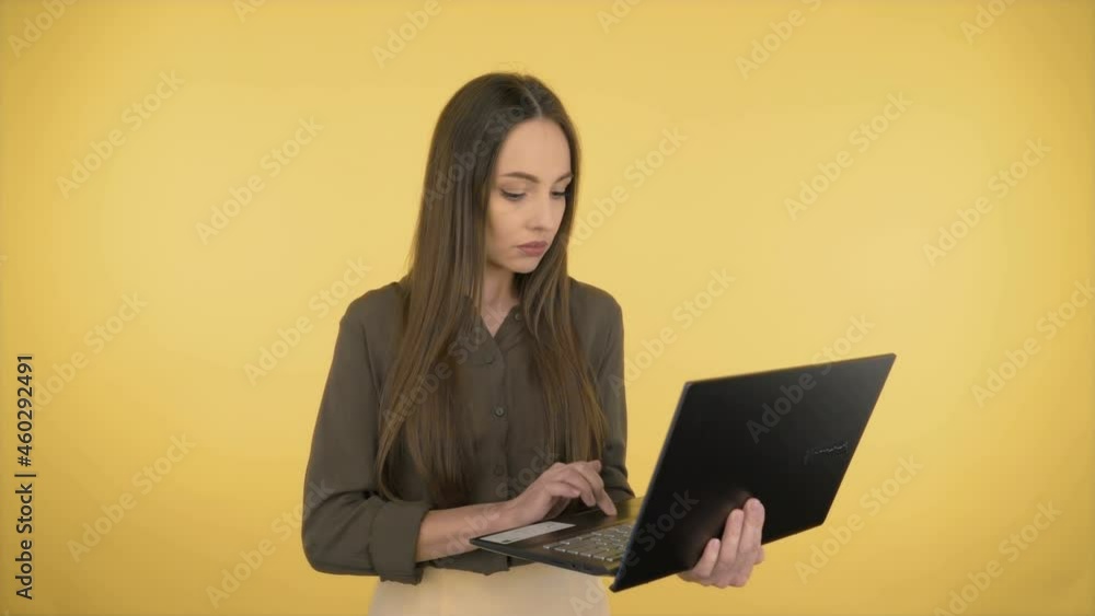 Attractive smiling business woman in a dark green office shirt. Woman working on a laptop while standing on yellow background. 4K Attractive smiling business woman in a dark green office shirt. Woman working on a laptop while standing on yellow background. 4K