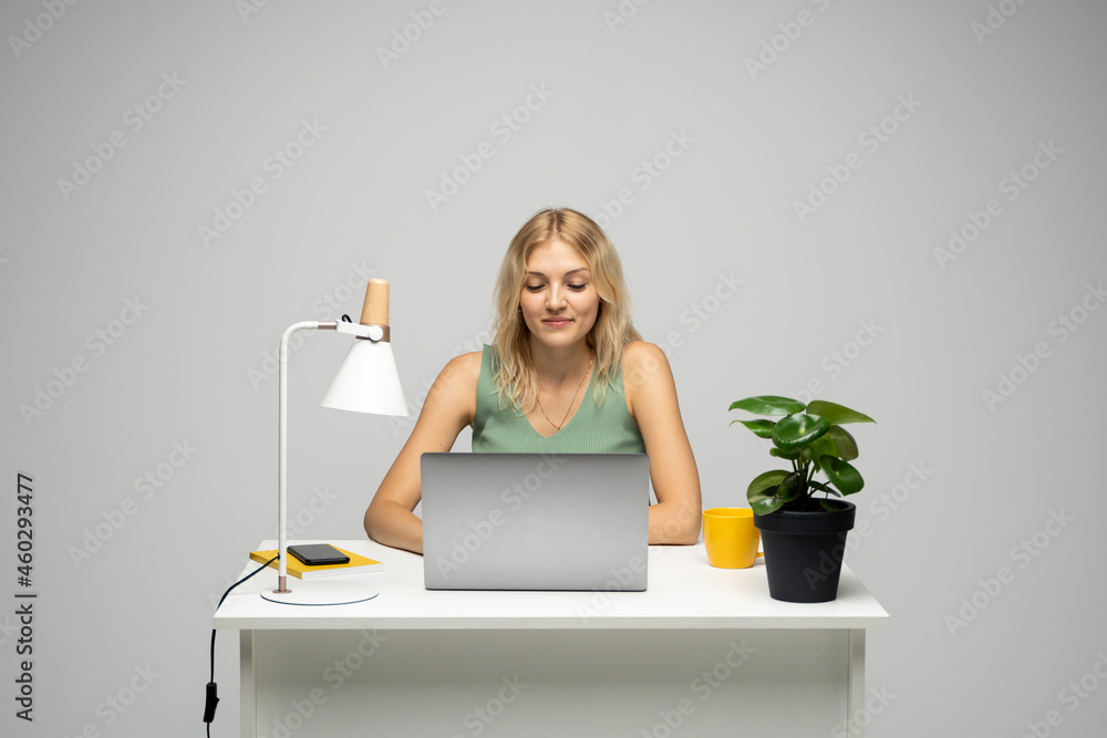 Portrait of a pretty young woman studying while sitting at the table with grey laptop computer, notebook. Smiling business woman working with a laptop isolated on a grey background. Portrait of a pretty young woman studying while sitting at the table with grey laptop computer, notebook. Smiling business woman working with a laptop isolated on a grey background.