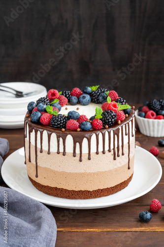 Three chocolate mousse cake with chocolate glaze, blueberries, raspberries and blackberries on top on a white plate on a dark wooden background. Cake with three layers of different taste. Copy space.