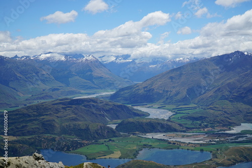 A landscape view of green fields and hills, river, Lake Wanaka and snowy mountain peaks from Roys Peak, Wanaka, South Island, New Zealand