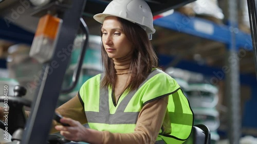 Live camera follows confident female driver autoloader operator sitting in warehouse forklift. Serious professional expert woman in hard hat operating machinery in industrial factory storage photo